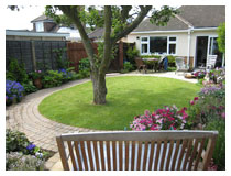 Oval lawn with curved path, patio laid in sandstone, borders and apple tree, seen from sitting-area.  Garden design and landscape gardening in Bedfordshire.